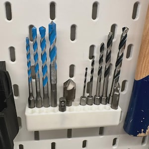 Può includere: Un organizer in plastica bianca montato su un pannello forato, contenente un set di punte da trapano. Il lato sinistro presenta quattro punte con spirali blu, mentre il lato destro contiene cinque punte in metallo nero, una svasatrice e un adattatore.