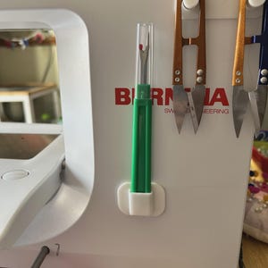 Peut inclure: Une machine à coudre blanche portant l'inscription rouge BERNINA SWISS ENGINEERING. Sur la façade sont fixés deux coupe-fils en métal cuivré et un découd-vite vert avec un capuchon en plastique transparent, maintenus par des supports blancs.
