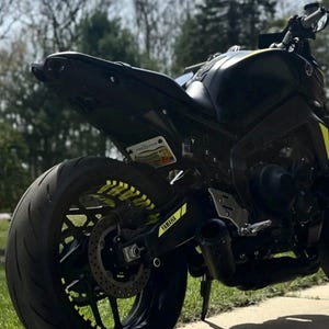May include: A black Yamaha motorcycle with yellow accents. The rear tire has yellow rim stickers. The exhaust pipe has the Yamaha logo. The motorcycle is parked on a concrete surface with grass in the foreground and trees in the background.