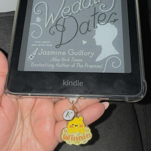 May include: A yellow Winnie the Pooh character keychain with a gold chain and a letter K charm. The keychain is attached to a Kindle e-reader with the book "Wedding Date" by Jasmine Guillory on the screen. The text on the screen reads "Love wasn't part of the deal. Wedding Date. Jasmine Guillory. New York Times Bestselling Author of The Proposal. Kindle."