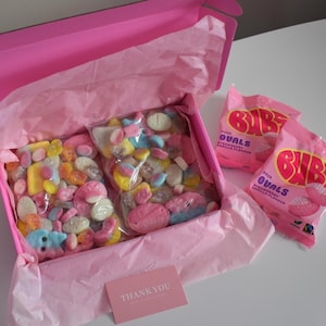 Puede incluir: Una caja de regalo rosa llena de caramelos de colores en bolsas de plástico transparente, forrada con papel de seda rosa. Se ven dos bolsas de caramelos Bubs Sour Ovals. También hay una pequeña tarjeta rosa con "Thank you".