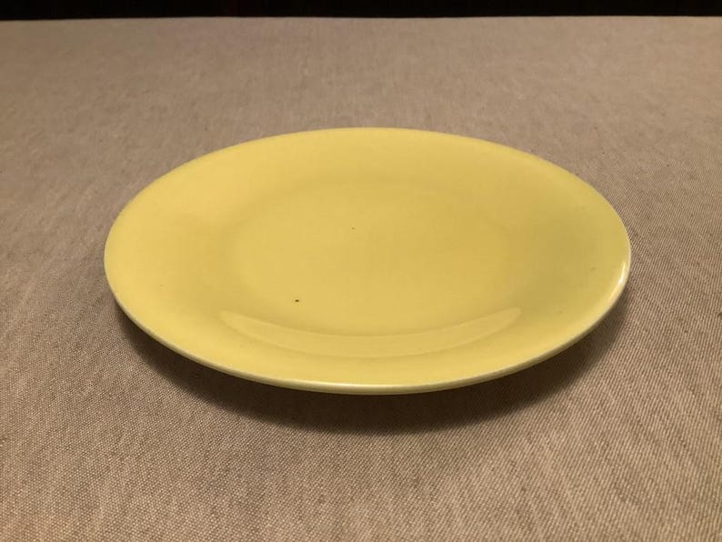Pu&ograve; includere: Un piatto rotondo giallo chiaro con un bordo leggermente rialzato. Il piatto &egrave; di un colore giallo pallido uniforme e sembra essere fatto di ceramica o materiale simile. &Egrave; centrato su una superficie dai toni neutri.