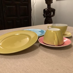 Pu&ograve; includere: Una collezione di stoviglie in ceramica dai colori pastello, tra cui un piatto ovale giallo, un piatto azzurro e un piatto rosa con una tazza e un piattino gialli. Gli articoli sono disposti su una tovaglia beige.