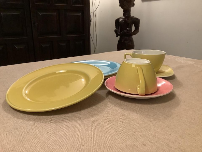 Pu&ograve; includere: Una collezione di stoviglie in ceramica dai colori pastello, tra cui un piatto ovale giallo, un piatto azzurro e un piatto rosa con una tazza e un piattino gialli. Gli articoli sono disposti su una tovaglia beige.
