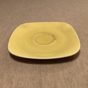 Pu&ograve; includere: Un piattino giallo pallido, di forma quadrata con angoli arrotondati. Il centro ha un'area incassata per una tazza. Il piatto in ceramica ha un aspetto leggermente usurato, suggerendo che sia vintage o ben usato. Il piattino &egrave; un pezzo semplice e funzionale.