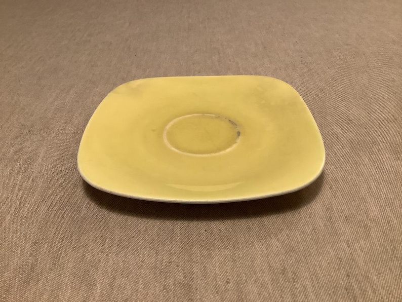 Pu&ograve; includere: Un piattino giallo pallido, di forma quadrata con angoli arrotondati. Il centro ha un'area incassata per una tazza. Il piatto in ceramica ha un aspetto leggermente usurato, suggerendo che sia vintage o ben usato. Il piattino &egrave; un pezzo semplice e funzionale.
