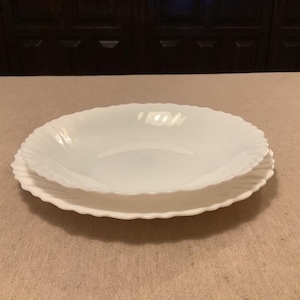 Puede incluir: Dos fuentes blancas con bordes festoneados apiladas sobre una superficie beige. El plato superior es un cuenco y el inferior es un plato. Los platos son de un material brillante y opaco, probablemente cerámica o porcelana.