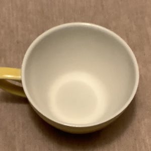 Pu&ograve; includere: Una tazza da t&egrave; in ceramica bianca con manico giallo. La tazza ha un'ampia apertura e una base arrotondata. L'interno &egrave; bianco e l'esterno &egrave; giallo pallido. La tazza &egrave; appoggiata su una superficie marrone.