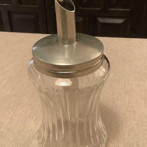 May include: A clear glass sugar dispenser with a silver lid and spout. The dispenser has vertical ridges and a rounded base.
