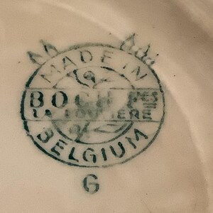 Pu&ograve; includere: Primo piano di un timbro in ceramica su una superficie bianca. Il timbro &egrave; circolare con la scritta "MADE IN BELGIUM" e "B.O. LA LOUVIERE" in blu scuro. La lettera "G" &egrave; sotto.