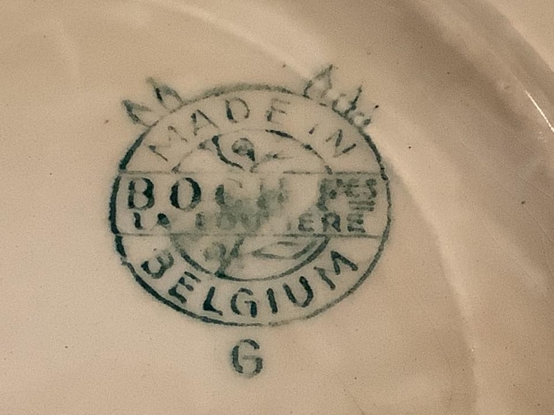 Pu&ograve; includere: Primo piano di un timbro in ceramica su una superficie bianca. Il timbro &egrave; circolare con la scritta "MADE IN BELGIUM" e "B.O. LA LOUVIERE" in blu scuro. La lettera "G" &egrave; sotto.