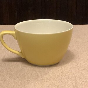 Pu&ograve; includere: Una tazza da t&egrave; in ceramica gialla con interno bianco. La tazza ha una forma arrotondata e un manico curvo. La tazza da t&egrave; &egrave; su una superficie di colore chiaro, con uno sfondo marrone scuro.