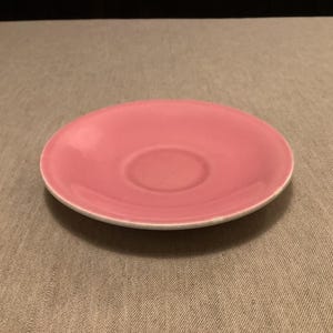Pu&ograve; includere: Un piattino in ceramica rosa con bordo bianco. Il piattino ha un centro leggermente concavo, progettato per contenere una tazza da t&egrave; o da caff&egrave;. La superficie liscia riflette la luce, evidenziando il colore rosa tenue. Un pezzo semplice ed elegante per servire bevande.