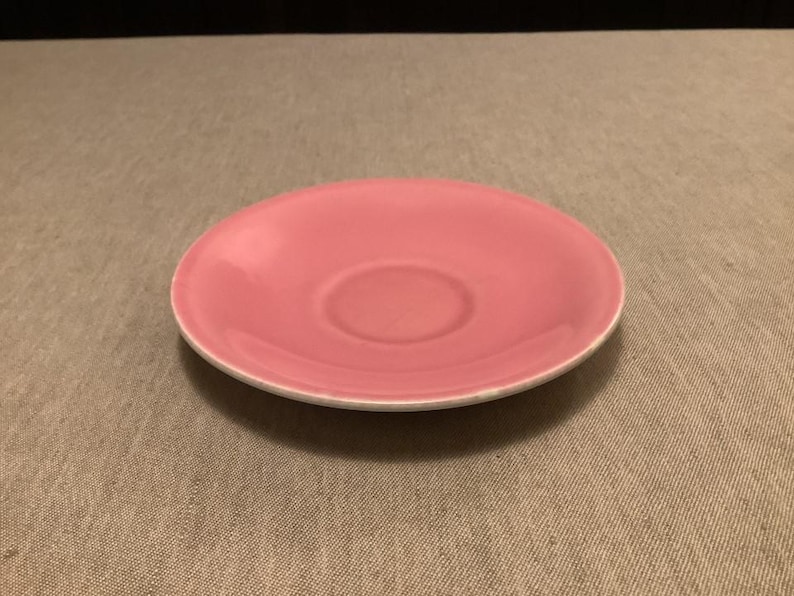 Pu&ograve; includere: Un piattino in ceramica rosa con bordo bianco. Il piattino ha un centro leggermente concavo, progettato per contenere una tazza da t&egrave; o da caff&egrave;. La superficie liscia riflette la luce, evidenziando il colore rosa tenue. Un pezzo semplice ed elegante per servire bevande.