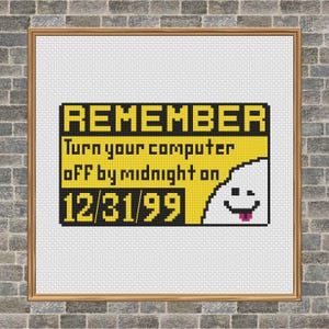 以下が含まれることがあります： 「REMEMBER Turn your computer off by midnight on 12/31/99」と書かれた黄色と黒の標識の額入りクロスステッチ。右側には笑顔の幽霊のような姿があります。フレームは明るい茶色です。