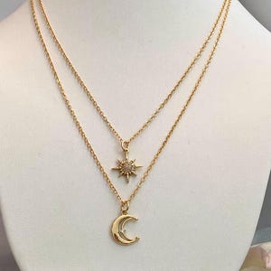 May include: A gold-colored layered necklace with celestial charms. The top layer features a sunburst pendant, while the bottom layer showcases a crescent moon pendant. The necklace is displayed on a white mannequin.
