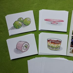 May include: A collection of watercolor illustrations on white cards, featuring food items like olives, a cake stand, and a layered dish. A "Horses" print and pens are also present, all arranged on a green surface.