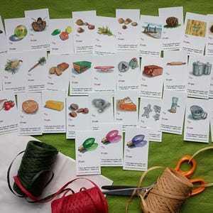May include: A collection of illustrated gift tags with various images, including a butterfly, fruit, and food items. The tags are arranged on a green surface with spools of twine, scissors, and a white bag.