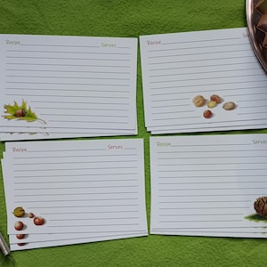 May include: A collection of lined recipe cards with decorative illustrations of nuts, leaves, and a pinecone. A wooden spoon and whisk are visible on the left, with a decorative metal plate in the background. The cards are on a green surface.