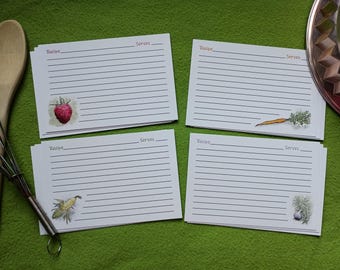 May include: Four white recipe cards with lined pages and illustrated accents of a raspberry, carrot, bird, and garlic. A wooden spoon and whisk are on the left, with a decorative metal plate on the right, all on a green surface.