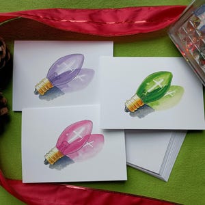 May include: Watercolor greeting cards featuring colorful Christmas light bulb designs in purple, green, and pink. Each card has a white background and a gold-colored base. The cards are arranged on a green surface with a red ribbon and pine cones.