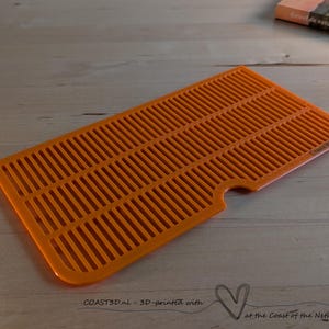 Pode incluir: Uma grelha retangular de plástico laranja com uma série de ranhuras paralelas e um pequeno recorte semicircular ao longo de uma das bordas mais longas. A superfície tem um acabamento texturizado e o texto COAST3D.nl está gravado.