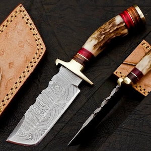 Puede incluir: Un cuchillo de caza con una hoja de acero Damasco y un mango de asta de ciervo con detalles rojos y marrones. El cuchillo se muestra con una funda de cuero.