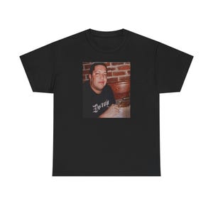 May include: Black t-shirt featuring a photograph of a person. The photograph is centered on the shirt and shows a person wearing a black shirt with the word "Detroit" in a stylized font. The background of the photograph is a brick wall.