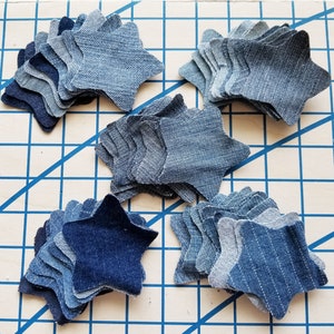 May include: A collection of six denim star shapes, each made of multiple layers of fabric. The stars are arranged in a pattern on a blue and white gridded surface.