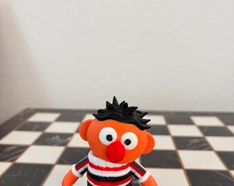 May include: A small 3D printed figurine of a character with orange skin, a round red nose, and black hair. The figure wears a shirt with horizontal red, white, and black stripes, black pants, and white shoes with red accents, standing in a upright position.