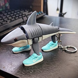 May include: A gray and white articulated shark figure with light blue sneakers for feet. The shark has a keychain attached. The sneakers have white soles and a white Nike swoosh.
