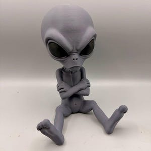 May include: A gray, 3D-printed alien figurine in a seated position with arms crossed over its chest. The figure features a large, smooth, oval-shaped head, prominent dark, almond-shaped eyes, and a small nose and mouth on a slender body.