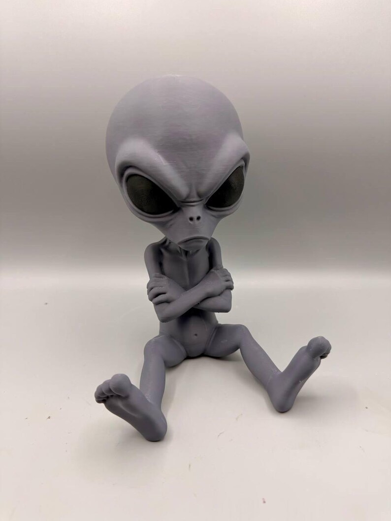 May include: A gray, 3D-printed alien figurine in a seated position with arms crossed over its chest. The figure features a large, smooth, oval-shaped head, prominent dark, almond-shaped eyes, and a small nose and mouth on a slender body.