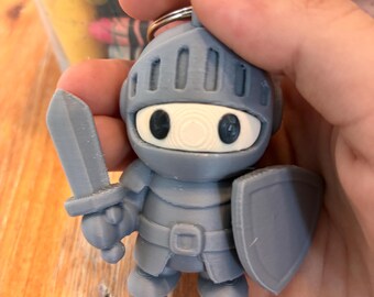 May include: A gray 3D-printed knight keychain featuring a helmet with a visor, a sword held in the right hand, and a shield held in the left hand. The knight has a white face plate with two dark circular eyes and a central circular nose detail.