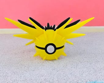 May include: A spherical yellow and black object resembling a creature with pointed, wing-like protrusions extending from the sides. The center features a circular white button surrounded by a black band, mimicking a classic ball design.