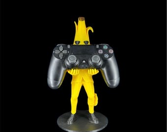 May include: A yellow banana-shaped figure wearing a suit and sunglasses, standing on a circular base. The figure holds a black video game controller with dual analog sticks, a directional pad, and four action buttons in its hands.