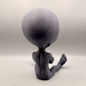 May include: A matte gray figurine of a seated humanoid creature with an oversized, smooth, rounded head, slender arms crossed over the chest, and elongated legs extending forward, showing visible spinal detail on the back.
