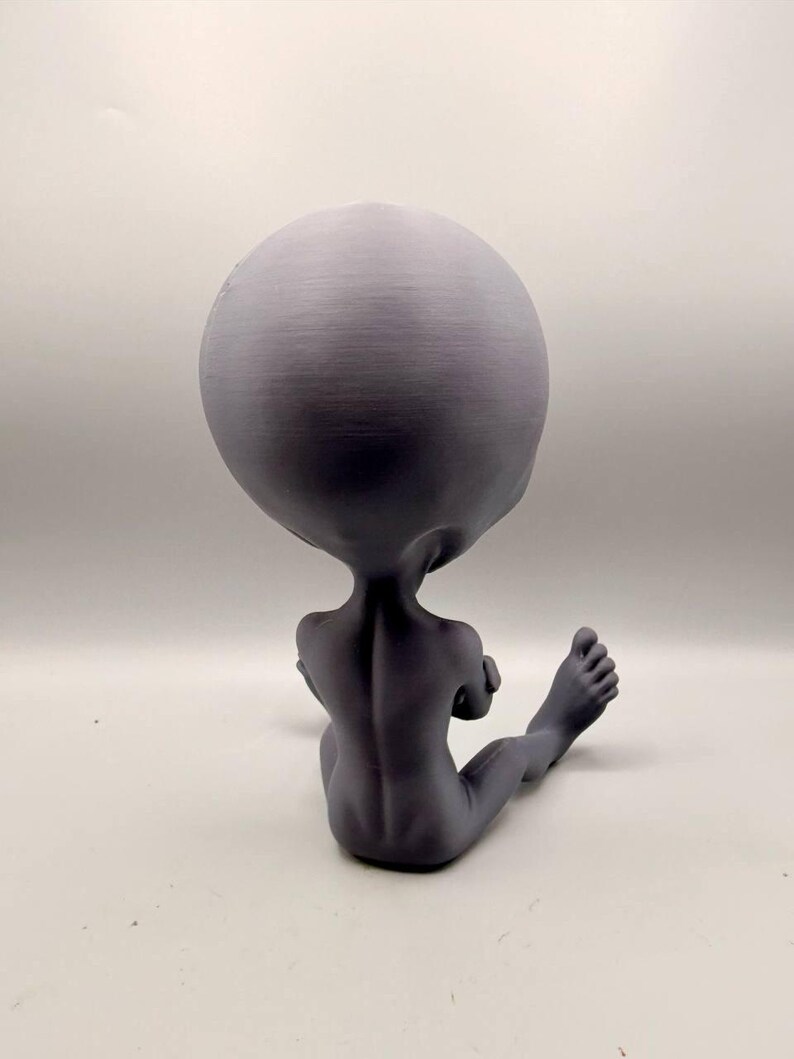 May include: A matte gray figurine of a seated humanoid creature with an oversized, smooth, rounded head, slender arms crossed over the chest, and elongated legs extending forward, showing visible spinal detail on the back.