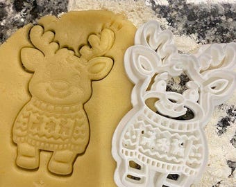 May include: A white plastic cookie cutter shaped like a reindeer wearing a patterned sweater, positioned next to a matching reindeer impression pressed into a flat piece of light tan cookie dough on a speckled surface.
