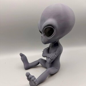 May include: A gray, 3D-printed alien figurine with a large, smooth, oval-shaped head and dark, recessed eyes. The figure is depicted in a seated position with its arms crossed over its chest and legs extended forward, showcasing a matte finish.