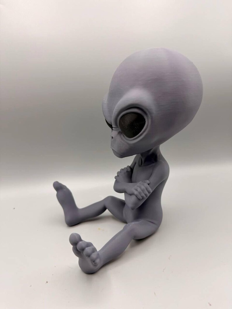 May include: A gray, 3D-printed alien figurine with a large, smooth, oval-shaped head and dark, recessed eyes. The figure is depicted in a seated position with its arms crossed over its chest and legs extended forward, showcasing a matte finish.