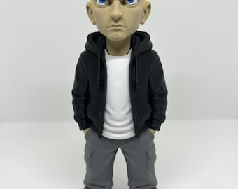 May include: A stylized figurine of a person with short, bright yellow hair and blue eyes. The figure wears a black hooded sweatshirt over a plain white t-shirt, paired with grey cargo pants and white sneakers. The hands are tucked into the pockets.