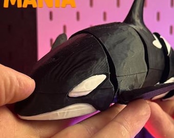 May include: A black and white articulated orca whale figurine featuring a segmented body design. The text Flexi MANIA is printed in blue and orange in the upper left corner. The toy has a smooth finish with visible joints between the sections.