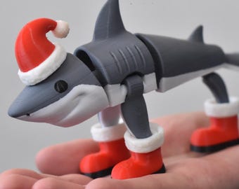 May include: A gray shark figurine with a white underbelly, wearing a red and white Santa hat on its head and red boots with white trim on its feet. The shark has a segmented body design with visible joints, a dorsal fin, and a tail fin.