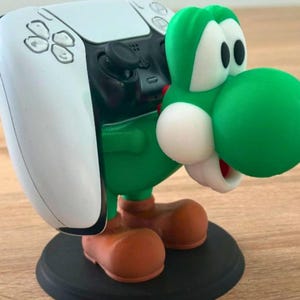 May include: A green and white character figurine acting as a controller stand, featuring a large green head with white cheeks and eyes, brown shoes, and a black circular base, supporting a white and black gaming controller on its back.