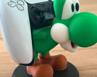 May include: A green and white character figurine acting as a controller stand, featuring a large green head with white cheeks and eyes, brown shoes, and a black circular base, supporting a white and black gaming controller on its back.