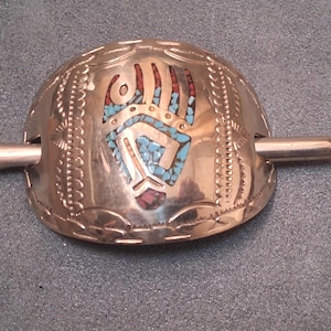 May include: A silver-toned hair barrette with a central design featuring a hand motif inlaid with red and turquoise stones. The barrette has a decorative edge and a silver pin for securing the hair accessory.