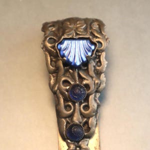 Puede incluir: Un marcapáginas de metal plateado con un diseño de vidrio azul. El marcapáginas presenta un patrón floral decorativo y tres acentos de vidrio azul.