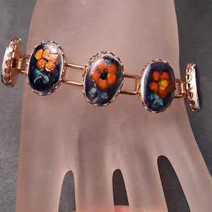 May include: A link bracelet featuring five oval-shaped metal settings with scalloped edges. Each setting contains a dark blue background with a hand-painted orange flower and green leaves. The links connecting the oval pieces are made of gold-tone metal.