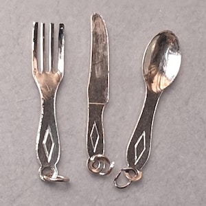 May include: Silver-colored miniature fork, knife, and spoon set. Each piece has a decorative diamond shape on the handle and a small loop at the end. The set is designed for a dollhouse or miniature display.